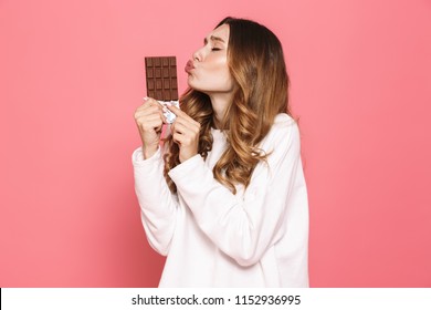Portrait of a happy young woman kissing chocolate bar isolated over pink background