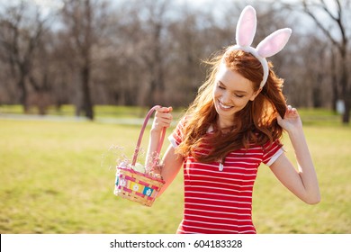 Portrait of a happy young woman with long red hair holding basket with painted eggs and celebrating easter holiday outdoors