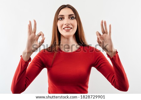 Portrait of a happy young woman with long chestnut hair showing OK gesture. Studio shot, white background. Human emotions, facial expression concept