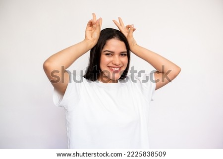 Portrait of happy young woman making bunny ears over head. Caucasian lady wearing white T-shirt looking at camera and smiling over white background. Humor and fun concept