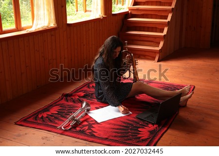 Portrait of a happy young woman with musical instrument and laptop.Girl with long hair composing music on a white sheet sitting on the floor holding a trumpet with bare legs in a wooden country house