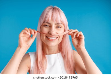 Portrait of happy young woman with natural long pink dyed hair holding a strand of hair as a moustache and smiling at camera, posing isolated over blue studio background. Beauty, hair care concept