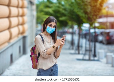 Portrait Of A Happy Young Woman Outdoors Wearing A Facemask While Texting On Her Cell Phone - Pandemic Lifestyle Concepts. Young Woman Wearing A Face Mask