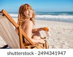 Portrait of happy young woman relaxing on wooden deck chair at tropical beach while looking at camera. Smiling girl with red hair and freckles enjoying vacation at seaside. Beautiful woman sunbathing.