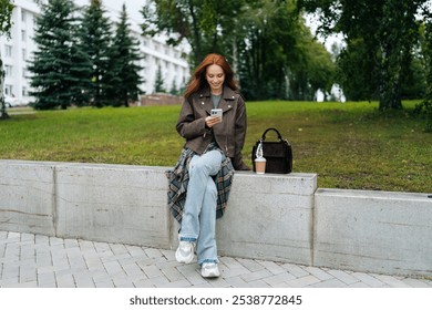 Portrait of happy young woman sitting on a bench enjoying beautiful day at park, using smartphone and drinking coffee during break. Stylish redhead woman relax alone, enjoying free time in outdoors. - Powered by Shutterstock