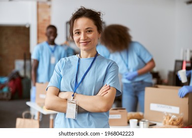 Portrait of happy young woman, volunteer in blue uniform smiling at camera while standing with arms crossed indoors. Team sorting, packing items in the background - Shutterstock ID 1968042223