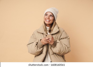Portrait of happy young woman in winter coat laughing and using cellphone isolated over beige background
