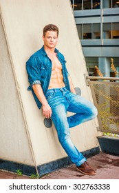Portrait of hard worker. Wearing Denim shirt, unbuttoned, sleeves rolled, jeans, leather shoes, a young, muscular, sexy guy casually standing against wall on balcony, bending leg, relaxing, thinking. - Shutterstock ID 302763338
