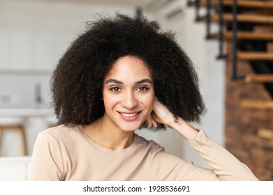 Portrait headshot of young pretty African American ethnic woman female freelancer with Afro curly hairstyle and beautiful smile, placed hand on cheek, looking at camera while standing in living room