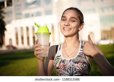 Portrait Of A Healthy Beautiful Cheerful Sports Fitness Slim Woman In Top With Tropical Print Holding Bottle With Water Outdoors In The Park On Grass With Sundown Light Looking Camera Make Thumbs Up.