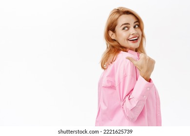 Portrait of healthy and happy young woman, pointing left and looking behind her shoulder, smiling carefree, standing over white background - Shutterstock ID 2221625739