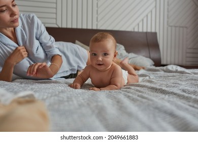 Portrait of healthy infant baby lying on tummy on bed next to his mom. Tummy time to help baby grow stronger and healthier. Young mother on bed near baby lying on belly, looking at camera.