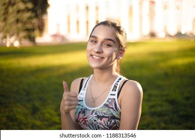Portrait Of A Healthy Smiling Beautiful Brunette Sports Fitness Slim Woman In Top With Tropical Print Outdoors In The Park On Grass With Sundown Light Looking Camera Showing Thumbs Up Gesture.