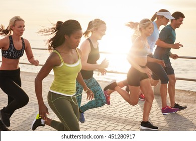 Portrait of healthy young men and women running race on seaside promenade. Group of young people sprinting outdoors at sunset.
