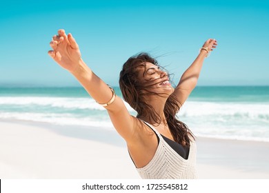 Portrait of healthy young woman standing on the beach with outstretched arms and feeling the breeze. Happy young woman feeling fresh and relaxing at ocean. Latin tanned woman with closed eyes.