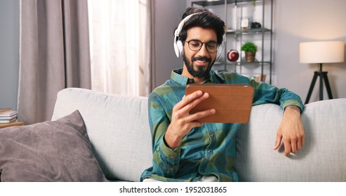 Portrait of Hindu young man watching movie or videos on tablet device in headphones while resting at home sitting in living room. Handsome bearded guy browsing online on gadget. Leisure concept - Powered by Shutterstock