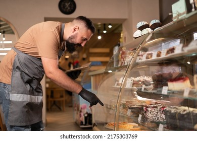 Portrait of a Hipster barista in apron using digital tablet standing near showcase at modern cafe. Smiling Indian bearded man receiving online orders with service mind. SME concept - Shutterstock ID 2175303769