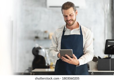 Portrait Of A Hipster Barista In Blue Apron Using Digital Tablet Standing At The Bar Of The Modern Cafes. Smiling Attractive Bearded Man Receiving Hot Drink Online Orders With Service Mind