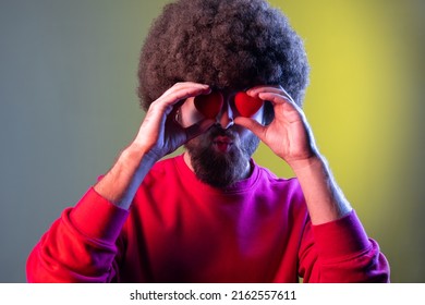 Portrait Of Hipster Man With Afro Hairstyle Covering Eyes With Small Red Hearts, Expressing Romantic Feelings, Wearing Red Sweatshirt. Indoor Studio Shot Isolated On Colorful Neon Light Background.