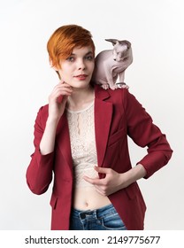 Portrait Of Hipster Redhead Young Woman With Short Hair, Dressed In Red Jacket, Blue Jeans And Playful Sphynx Cat Sitting On Her Shoulder. Front View Studio Shot On White Background. Part Of Series