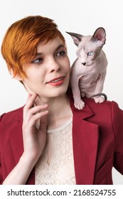 Portrait Of Hipster Redhead Young Woman With Short Hair, Dressed In Red Jacket And Playful Sphynx Cat Sitting On Her Shoulder. Studio Shot On White Background. Part Of Series.