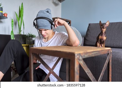 Portrait of hipster teenager in hat with headphones sitting on floor at home. Trendy handsome guy looking at smartphone screen, dog toy terrier looking at camera. Youth, lifestyle and leisure