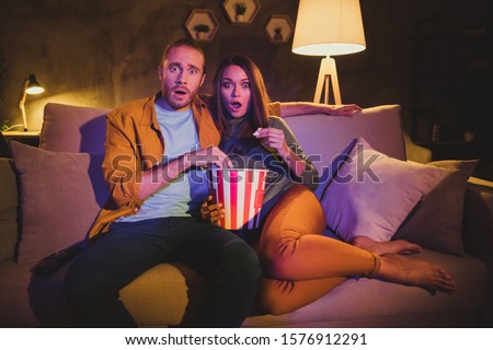 Portrait of his he her she nice attractive lovely scared afraid frightened married spouses sitting on divan watching horror genre film resting at home house apartment living-room indoors