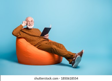 Portrait Of His He Nice Attractive Cheerful Cheery Glad Bearded Grey-haired Man Sitting In Bag Chair Reading Book Enjoying Rest Relax Pastime Touching Specs Isolated Over Blue Pastel Color Background