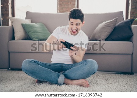 Portrait of his he nice attractive crazy addicted glad cheerful cheery guy sitting on carpet using cell playing game having fun at modern industrial loft brick interior style living-room