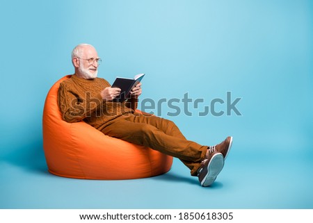 Portrait of his he nice attractive focused cheerful cheery wise smart clever bearded grey-haired man sitting in bag chair reading academic book isolated over blue pastel color background