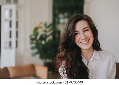 Portrait Of Hispanic Brunette Woman With Wavy Dark Hair Wearing White Shirt, Looking At Camera, Smiling At Home. Successful Entrepreneur Toothy Smiling, Happy To Develop Business Using New Strategies.