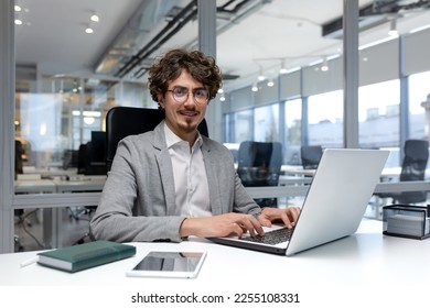 Portrait of hispanic businessman, man with curly hair and beard wearing glasses smiling and looking at camera, programmer typing on laptop inside bright office. - Shutterstock ID 2255108331