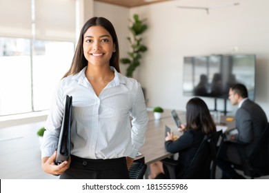 Portrait of hispanic businesswoman with a file standing in meeting room with colleagues disucssing in background