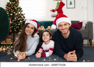 Portrait Of A Hispanic Family Of Three Lying Next To A Christmas Tree At Home And Looking Ready To Celebrate