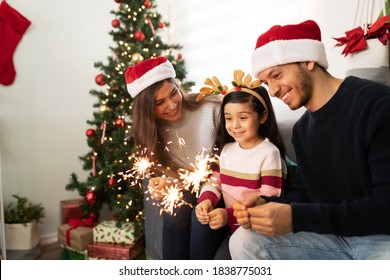 Portrait Of A Hispanic Family Using Sparklers At Home While A Little Girl Looks Very Impressed On Christmas