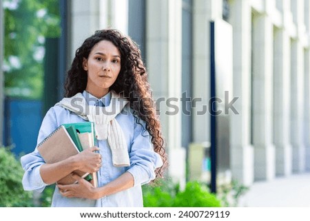 Portrait of a Hispanic female student standing on the street near an office building, holding textbooks and notebooks, looking at the camera.