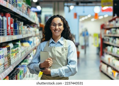 Portrait of a Hispanic female supermarket worker, saleswoman looking at the camera and smiling holding a tablet computer for product inspection, among racks and shelves in a store with goods. - Powered by Shutterstock