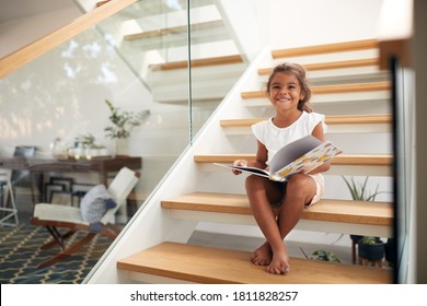 Portrait Of Hispanic Girl Sitting On Staircase In Modern Home Reading Book