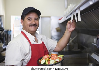 Portrait Of A Hispanic Male Waiter With Plate Of Food Standing In Restaurant Kitchen