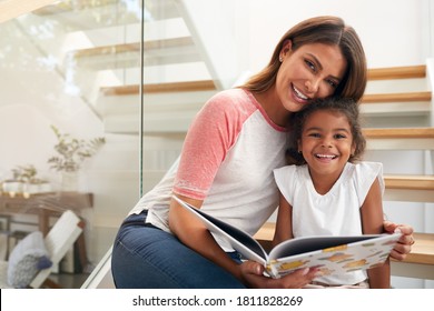 Portrait Of Hispanic Mother And Daughter Sitting On Staircase In Modern Home Reading Book Together