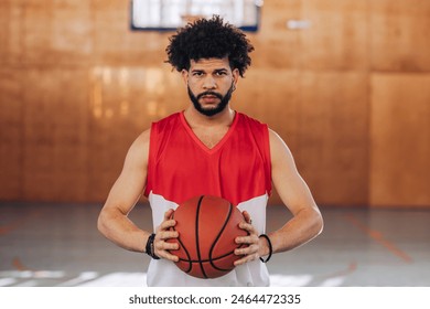 Portrait of a hispanic professional basketball player with the afro hair holding a ball with both hands while standing on an indoor court and looking at the camera. Sport, active lifestyle concept. - Powered by Shutterstock