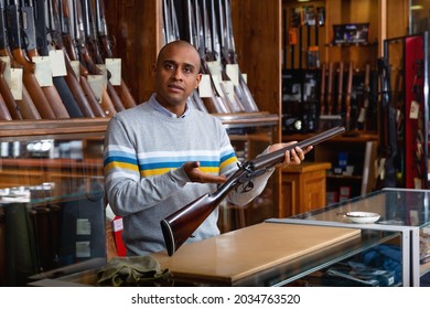 Portrait Of Hispanic Salesman Showing Collectible Old Rifled Musket On Background With Rack Full Of Vintage And Modern Firearms In Gun Store