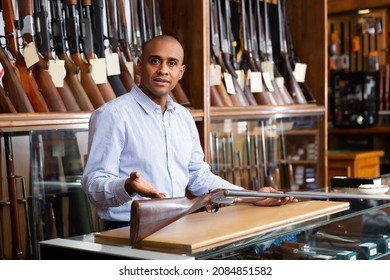 Portrait Of Hispanic Salesman Showing Collectible Old Rifled Musket On Background With Rack Full Of Vintage And Modern Firearms In Gun Store