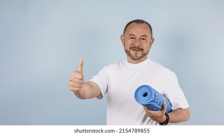 Portrait Of Hispanic Senior Man Holding Yoga Mat With Thumbs Up On Blue Background In Mexico Latin America