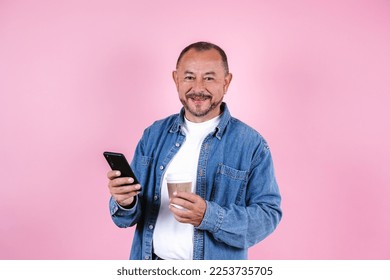 portrait of hispanic senior man wearing casual clothes and holding a mobile phone and coffee cup on blue background in Mexico Latin America - Powered by Shutterstock