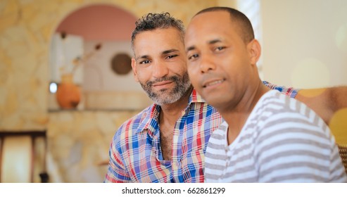 Portrait Of Homosexual Couple, Happy Gay People Smiling At Camera, Sitting On Sofa At Home. Same Sex Marriage Between Hispanic Men.