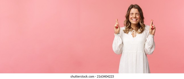 Portrait Of Hopeful Lucky Cute Blond Girl In White Dress Praying, Anticiapte Miracle, Making Wish To Win, Cross Fingers Good Luck, Smiling And Close Eyes And Pleading God, Pink Background