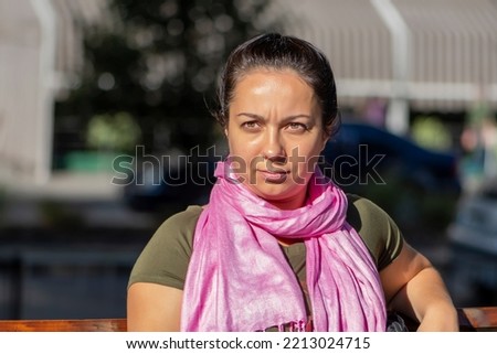 portrait of a horny 35-40 year old woman with a pink scarf around her neck, sitting on a bench against a neutral city background, sunny day, summer weather.