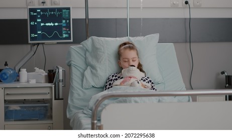 Portrait Of Hospitalized Sick Child Falling Asleep While Holding Teddy Bear In Hands During Medical Examination In Hospital Ward. Tired Kid Suffering Recovery Surgery Against Disease Infection