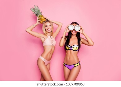 Portrait of hot, charming, stylish, pretty, trendy, crazy ladies, tourists in swim suits, blonde having ananas on her head, brunette having coconut on eyes' place, standing over pink background